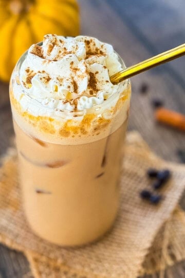 Glass of homemade iced pumpkin spice latte with ice cubes and whipped cream, surrounded by coffee beans and cinnamon sticks.