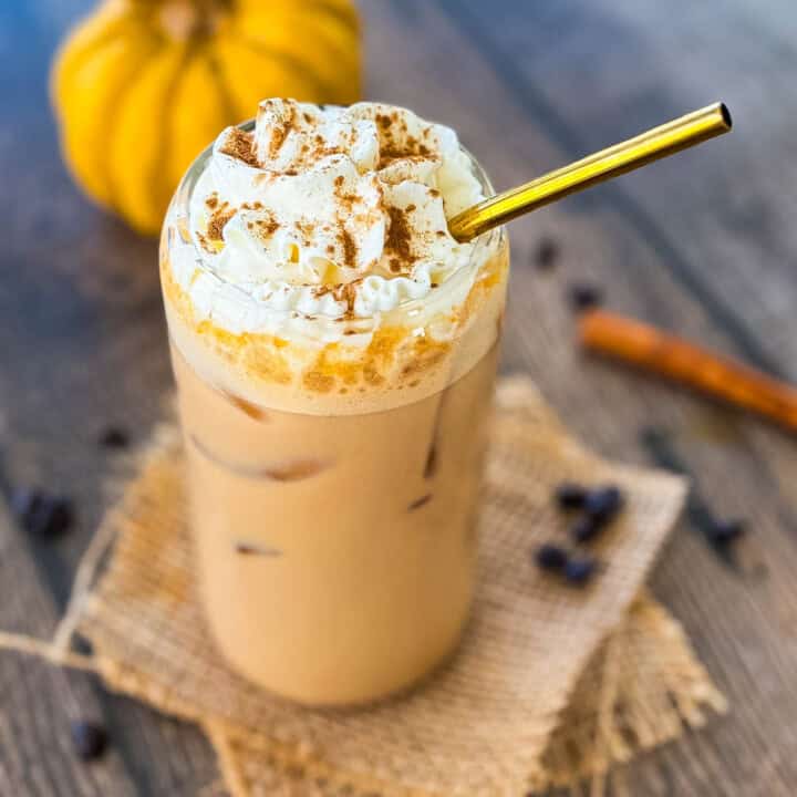 Glass of homemade iced pumpkin spice latte with ice cubes and whipped cream, surrounded by coffee beans and cinnamon sticks.