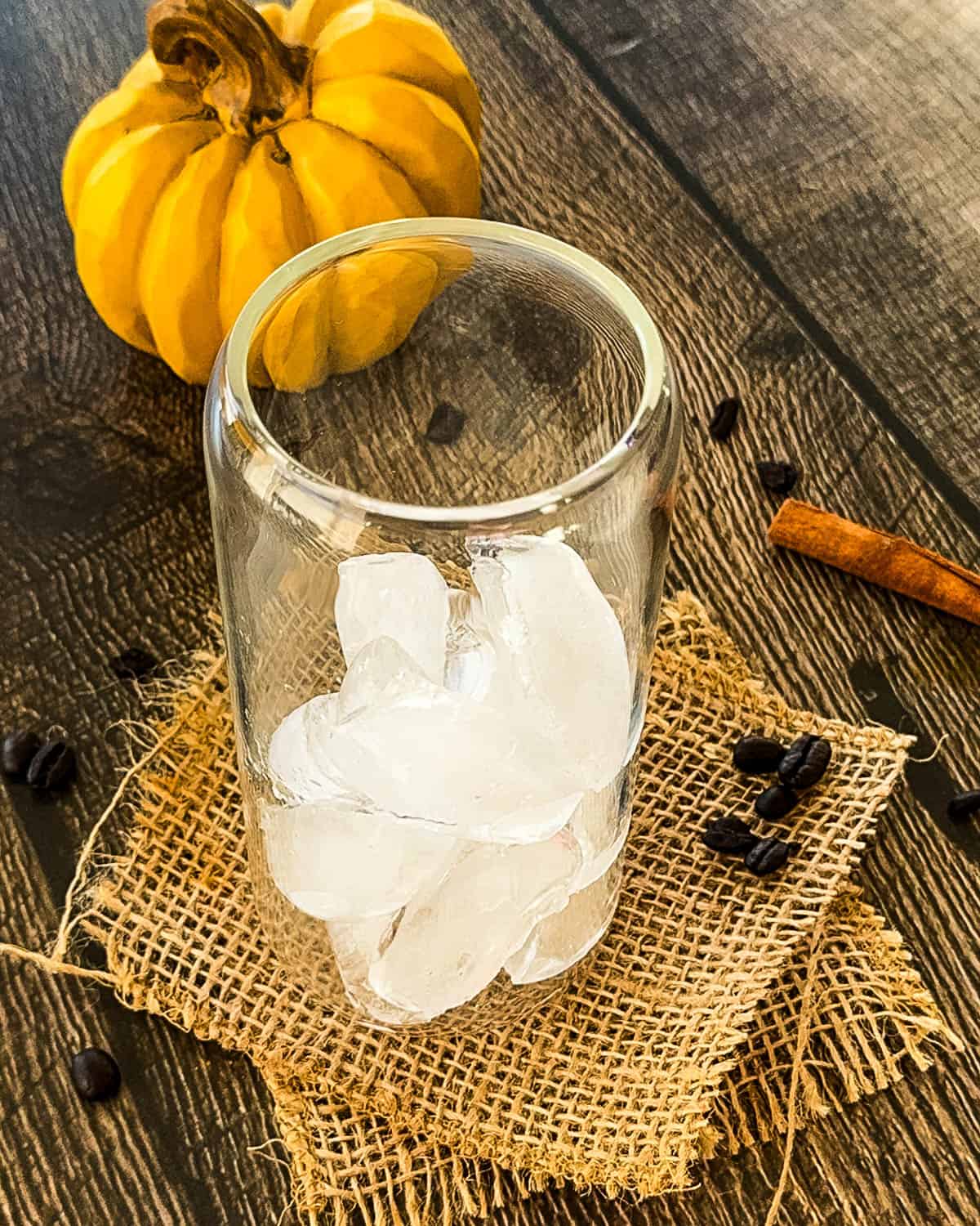 Tall glass filled with ice cubes on a rustic wooden table with a mini pumpkin, coffee beans, and cinnamon stick in the background.