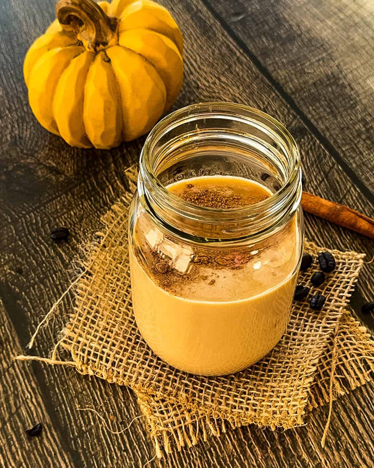 Mason jar filled with creamy pumpkin spice latte mixture sitting on a burlap square with fall decor and coffee beans scattered around.