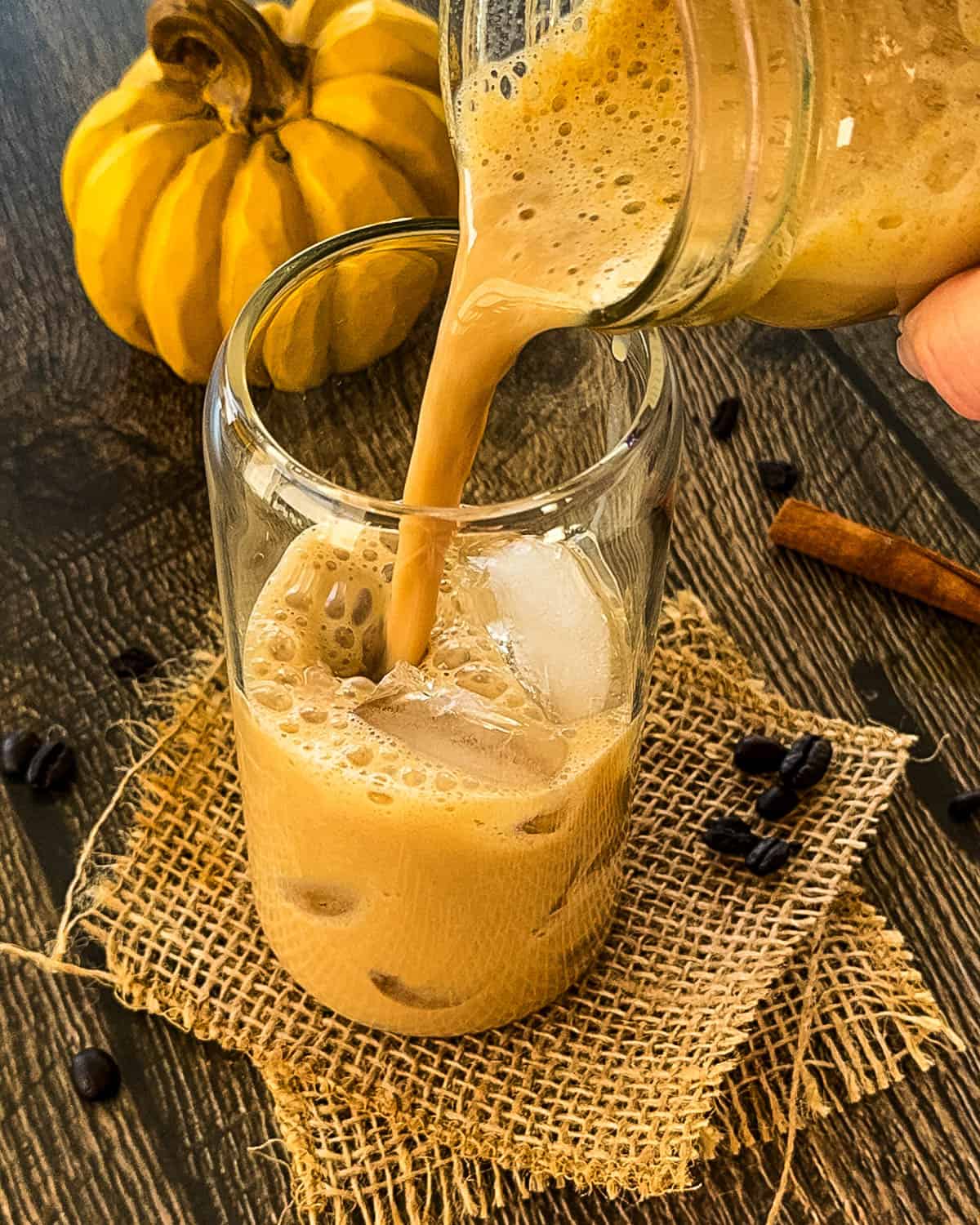 Pumpkin spice latte being poured from a mason jar into a tall glass of ice on a rustic wooden surface.