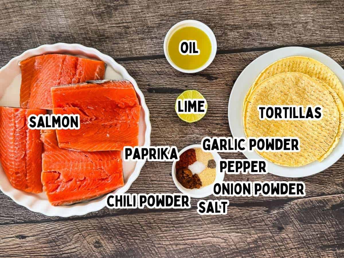 Ingredients for air fryer salmon tacos displayed on a wooden surface: raw salmon fillets, corn tortillas, olive oil, lime, and small bowls of chili powder, smoked paprika, garlic powder, onion powder, salt, and black pepper.