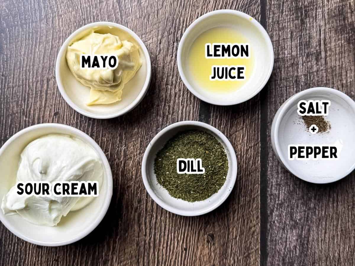 Small white bowls of mayonnaise, sour cream, dried dill, lemon juice, salt, and pepper arranged on a wooden surface, labeled with text.