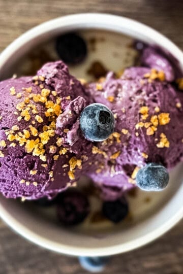 Scoops of Ninja Creami blueberry cheesecake ice cream in a white bowl topped with graham cracker crumbs and fresh blueberries.