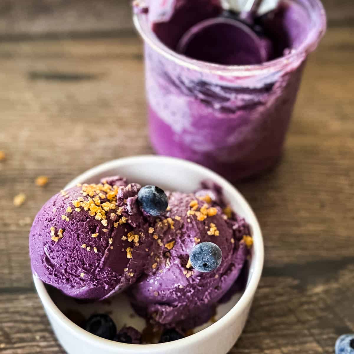 Scoops of Ninja Creami blueberry cheesecake ice cream in a white bowl topped with graham cracker crumbs and fresh blueberries.