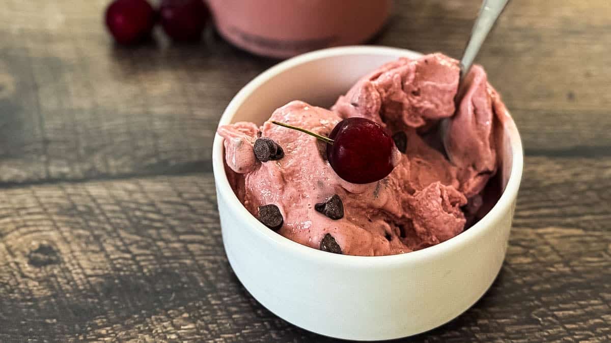 Bowl of homemade Ninja Creami cherry chocolate chip ice cream topped with a fresh cherry and chocolate chips on a rustic wooden table.