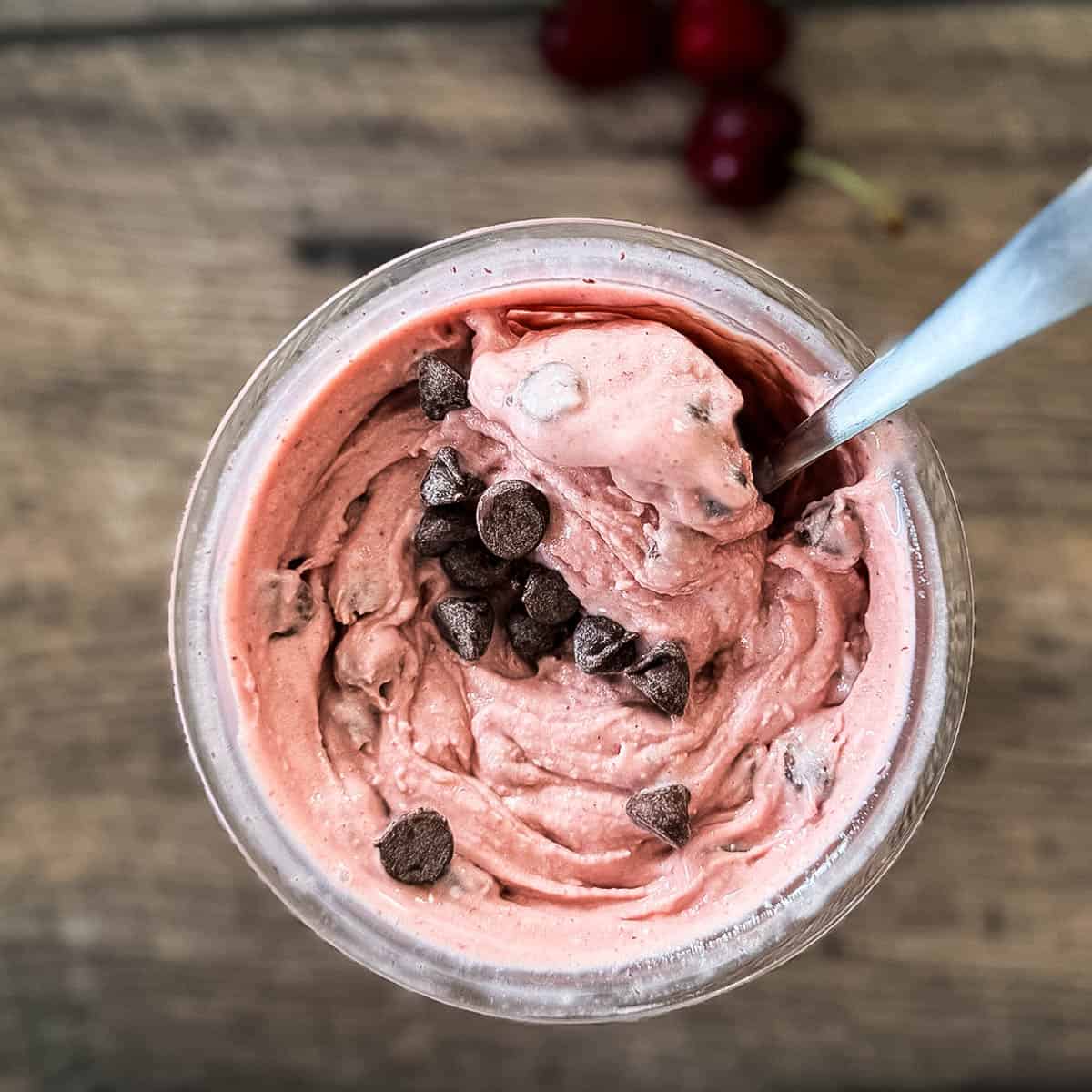 Creamy Ninja Creami cherry chocolate chip ice cream in a pint container, topped with chocolate chips, with fresh cherries in the background.
