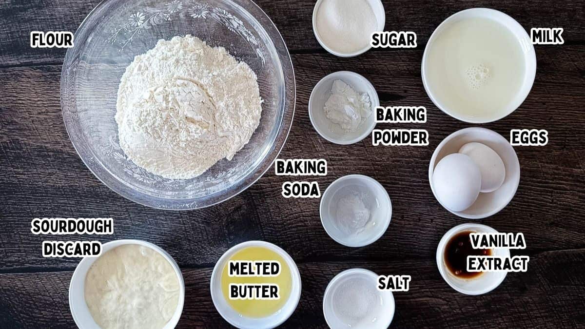 Labeled waffle ingredients in small bowls on a wooden surface, including flour, eggs, milk, sourdough discard, and vanilla.