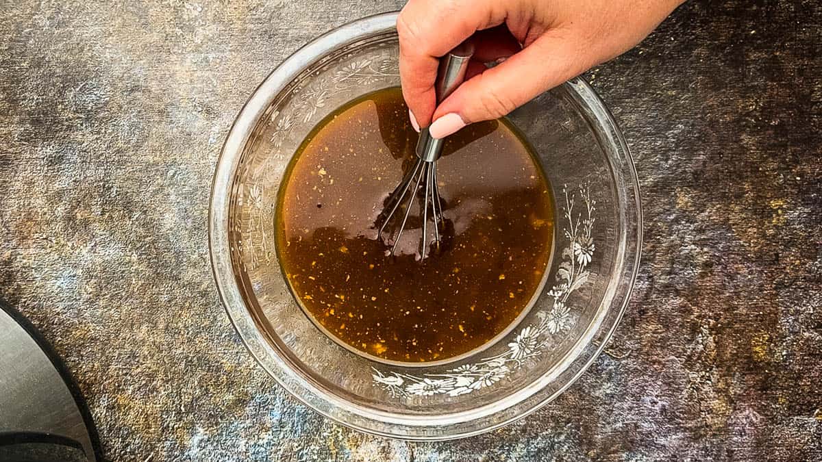 Hand whisking BBQ sauce, vinegar, broth, and sugar in a glass mixing bowl.