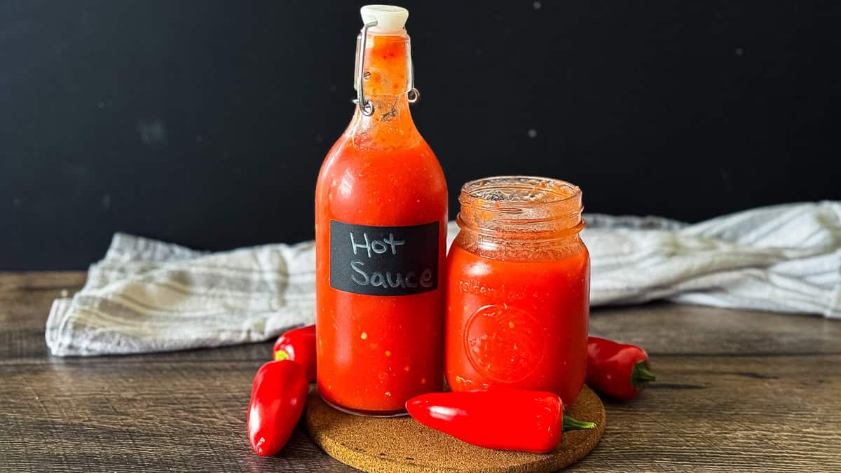 Bottle and jar of homemade hot sauce placed on a rustic wooden table with a striped towel in the background.