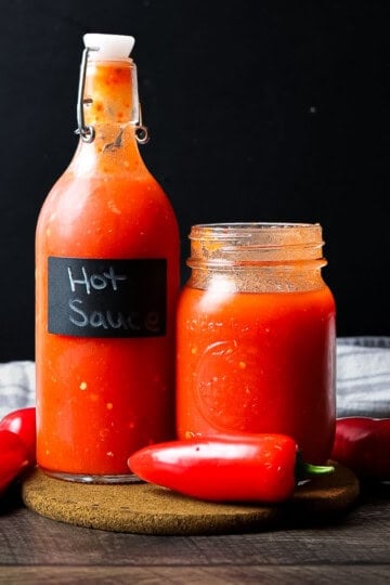 A glass swing-top bottle and a mason jar filled with bright red homemade hot sauce, surrounded by fresh red peppers.
