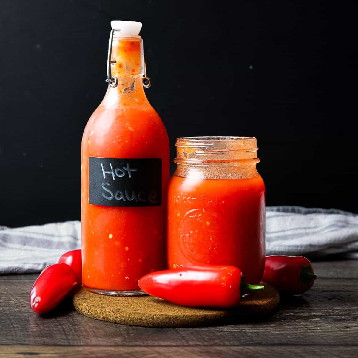 A glass swing-top bottle and a mason jar filled with bright red homemade hot sauce, surrounded by fresh red peppers.
