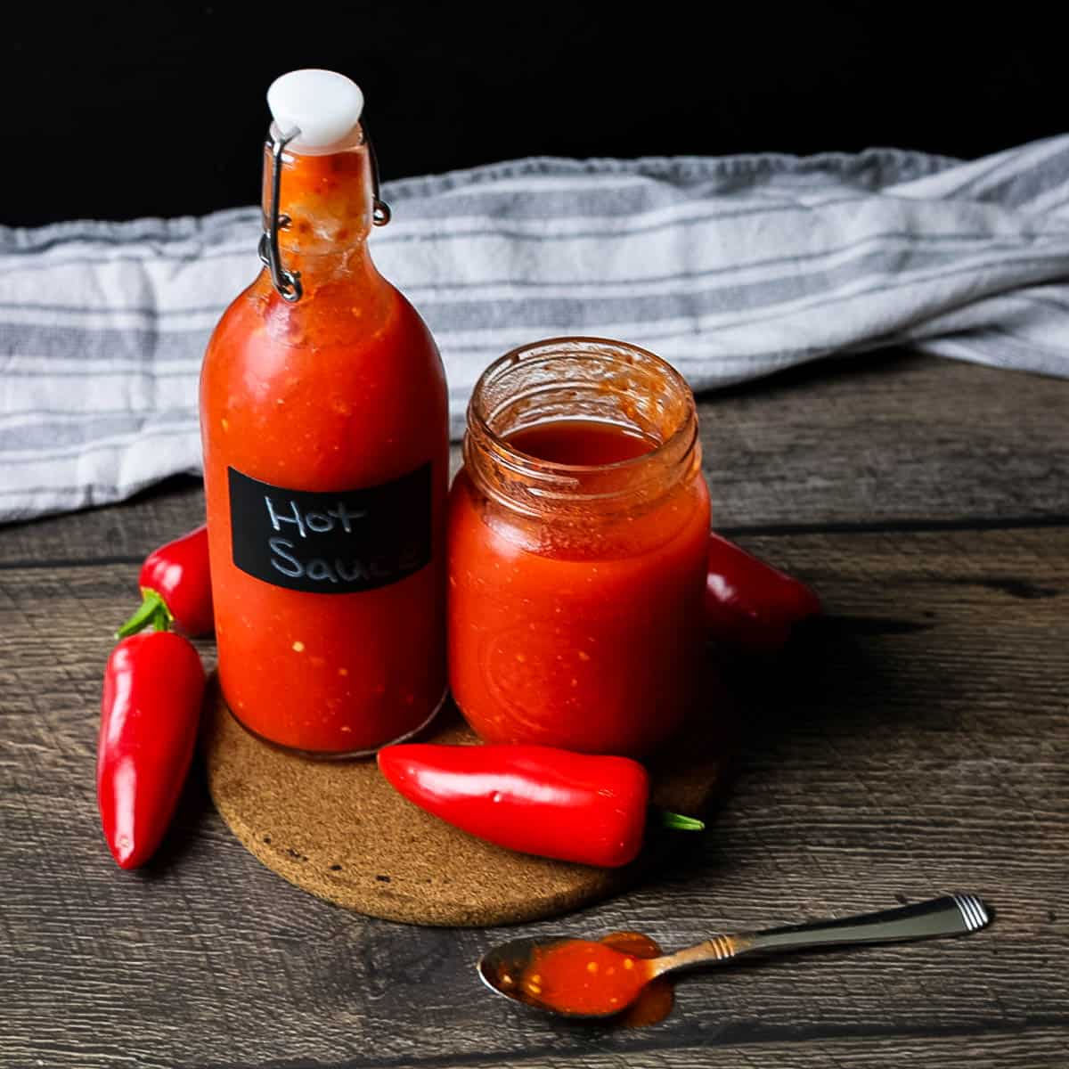 Jar and bottle of homemade hot sauce sitting on a cork trivet with fresh red peppers around them.