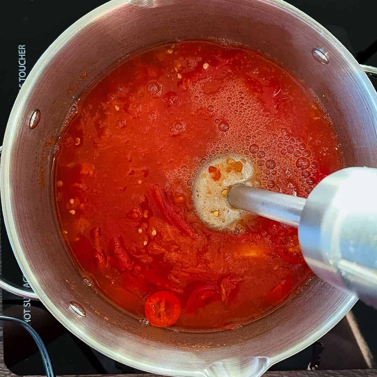 Immersion blender mixing cooked tomatoes, peppers, and garlic into a smooth hot sauce in a saucepan.