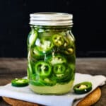 A glass mason jar filled with homemade refrigerator pickled jalapeños, sitting on a wooden board with fresh jalapeño slices scattered around.