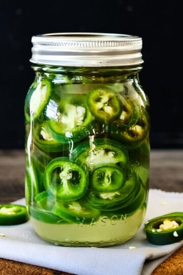 A glass mason jar filled with homemade refrigerator pickled jalapeños, sitting on a wooden board with fresh jalapeño slices scattered around.