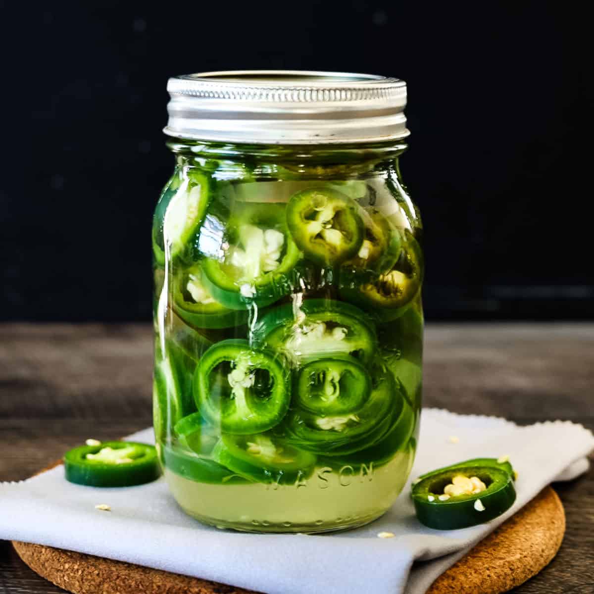 A glass mason jar filled with homemade refrigerator pickled jalapeños, sitting on a wooden board with fresh jalapeño slices scattered around.