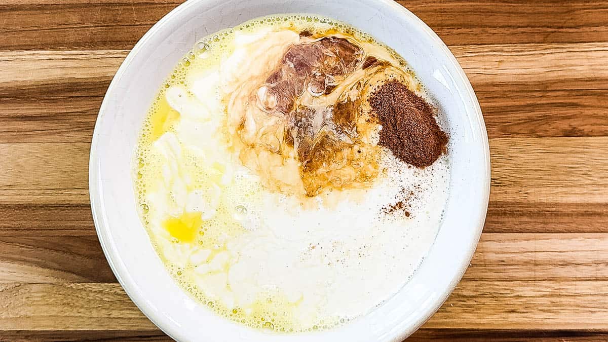 Creamy mixture with eggs, vanilla, cinnamon, and nutmeg in a white bowl on a wooden surface.