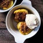 Two air fryer baked apple halves topped with golden oat crumble and a scoop of vanilla ice cream in a white ramekin on a wooden surface.