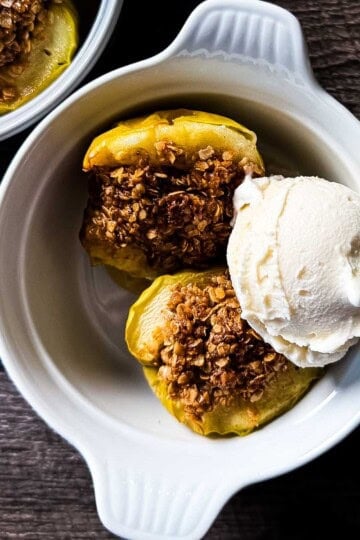 Two air fryer baked apple halves topped with golden oat crumble and a scoop of vanilla ice cream in a white ramekin on a wooden surface.