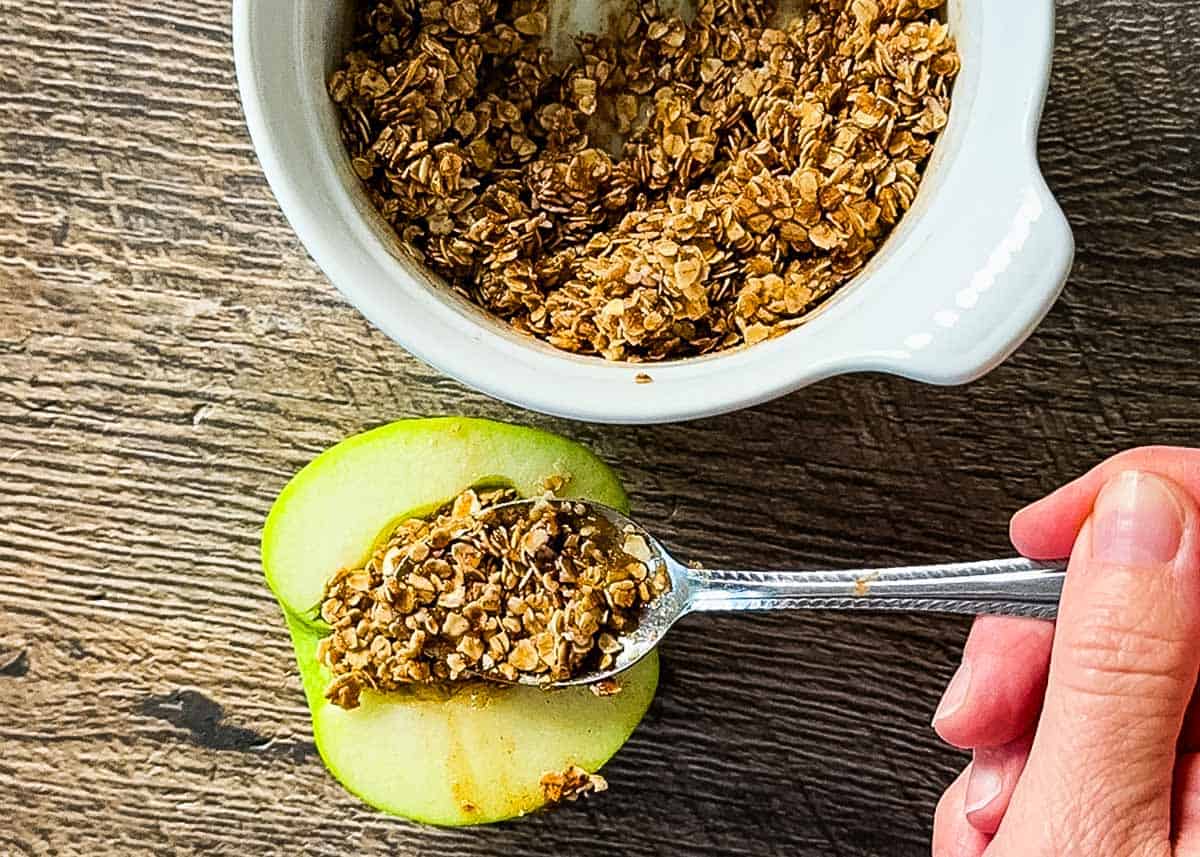 Hand spooning oat crumble mixture onto a cored green apple half with a white bowl in the background.