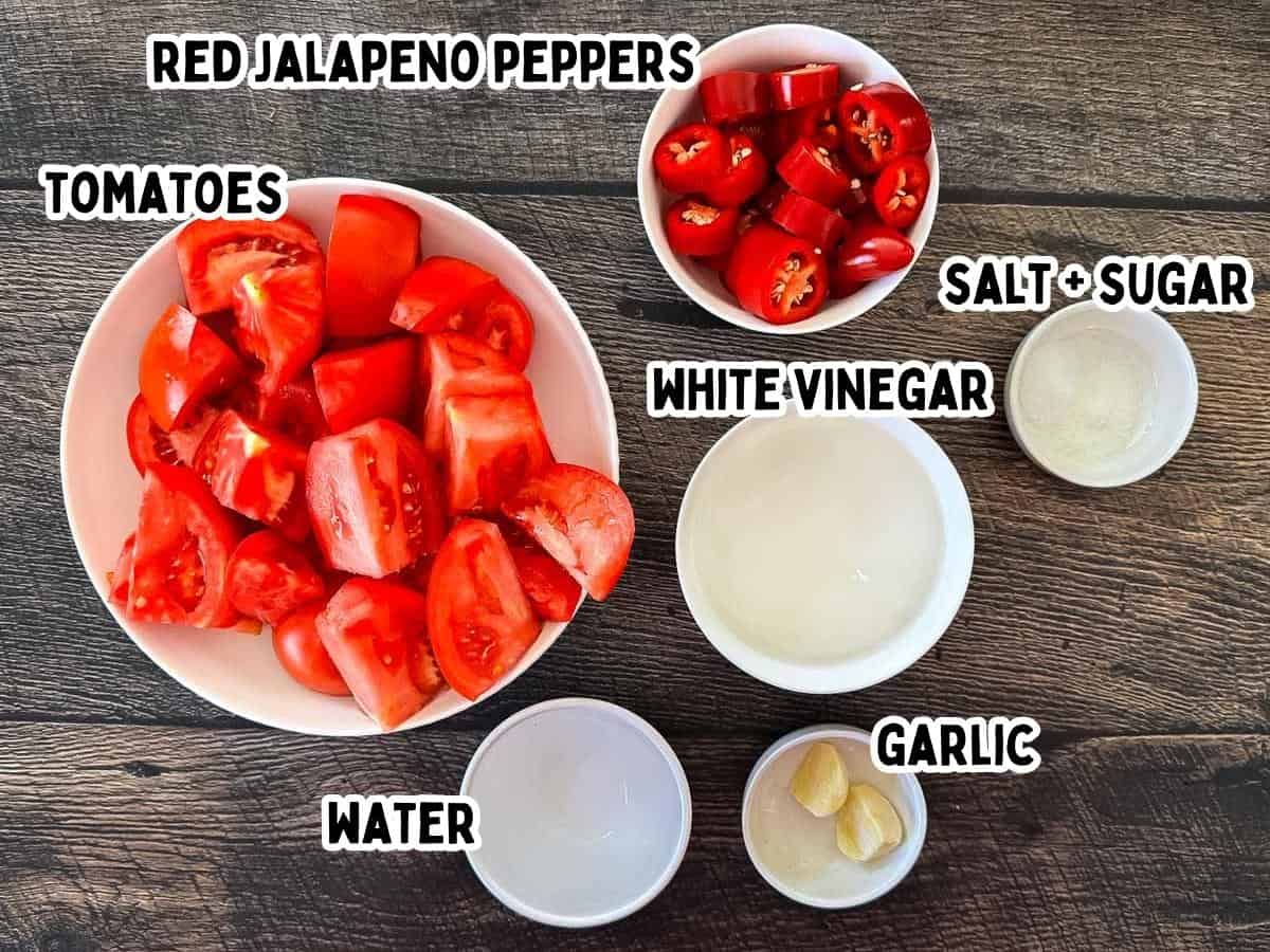 Fresh tomatoes, red jalapeños, garlic, vinegar, sugar, and salt arranged in small bowls on a wooden surface.