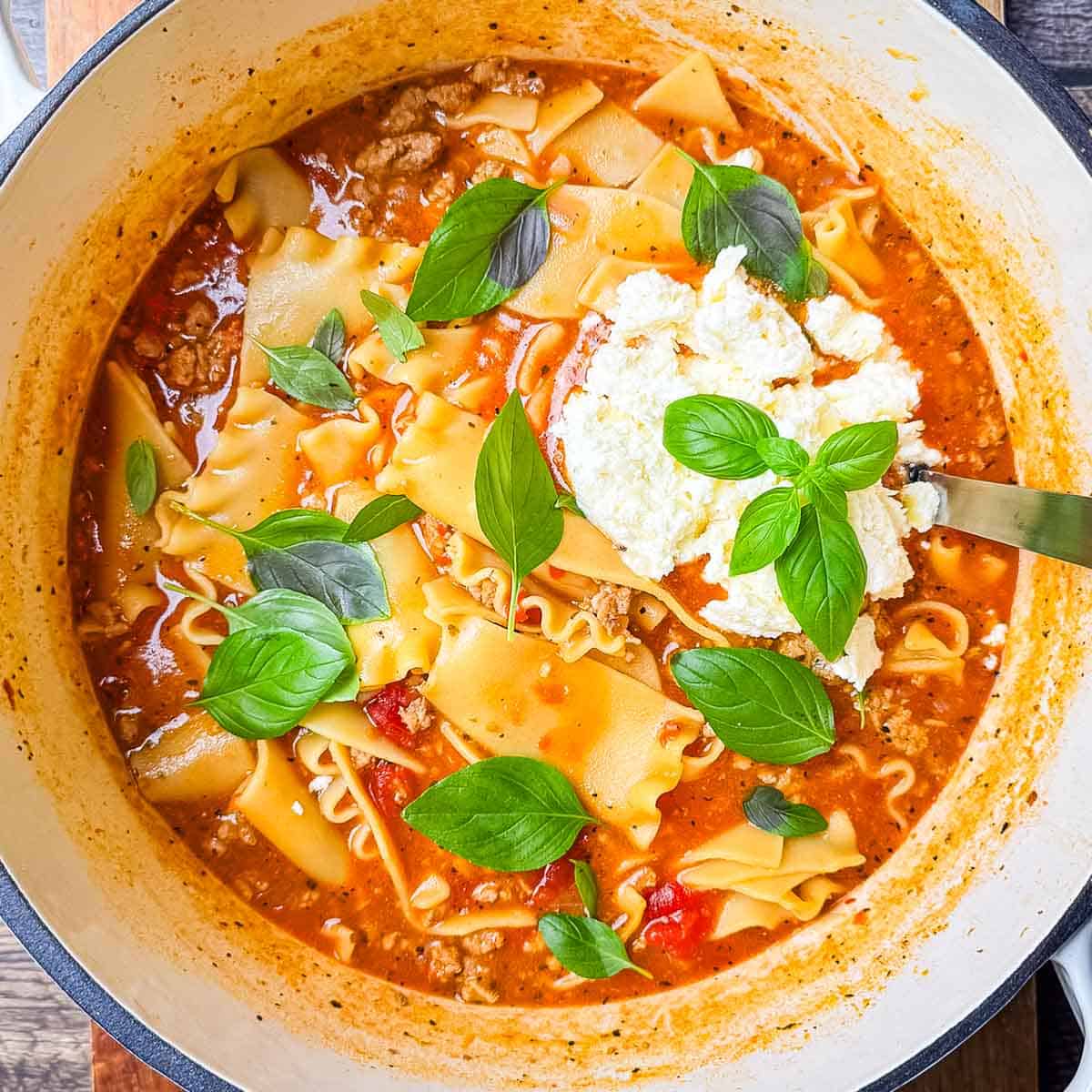 A Dutch oven filled with hearty lasagna soup topped with creamy cheese and fresh herbs.