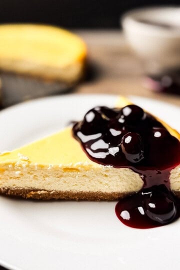A slice of smoked cheesecake on a white plate topped with glossy blueberry sauce, showing its creamy texture and buttery graham cracker crust.