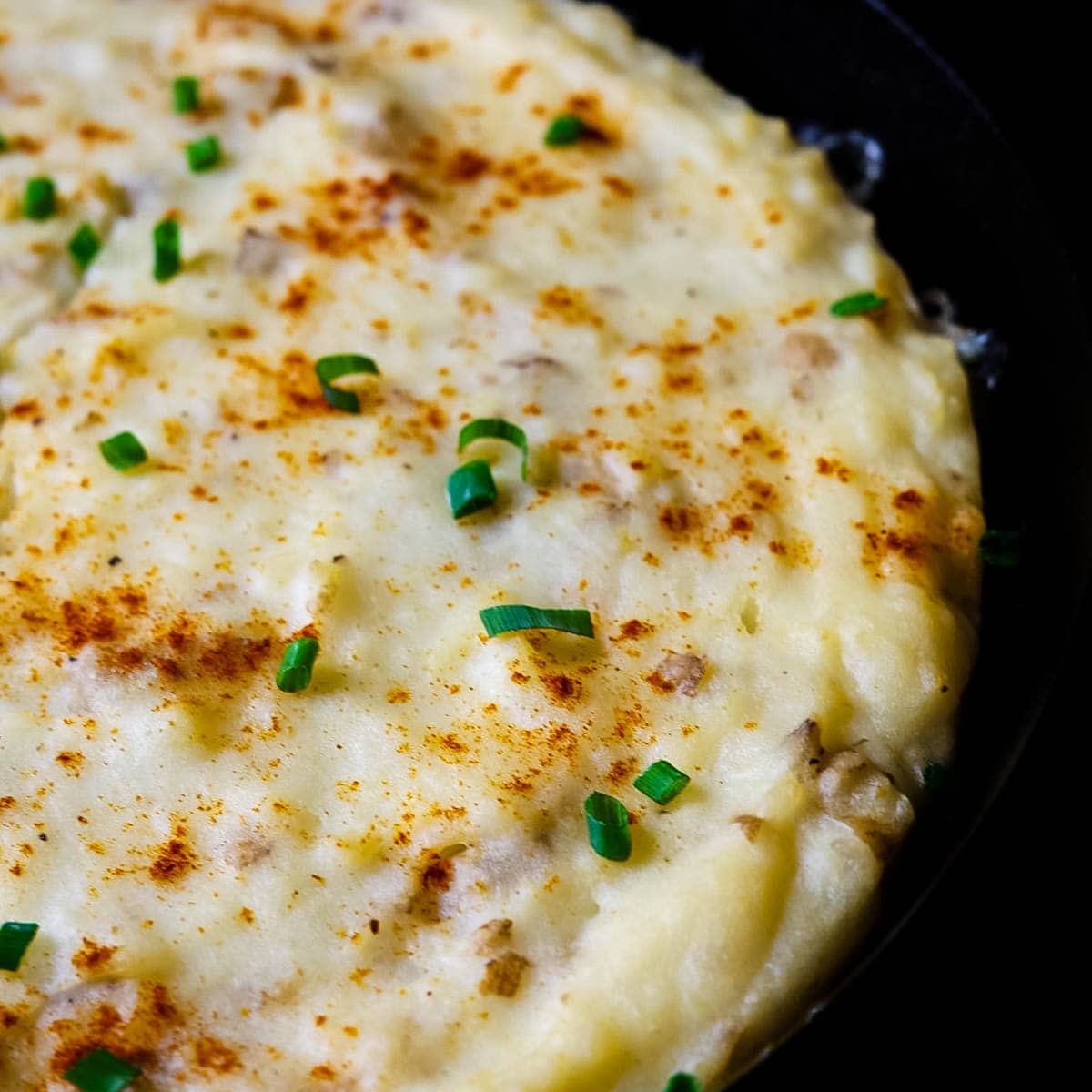 Angled shot of smoked mashed potatoes showing creamy texture with paprika and chopped chives on top in a cast iron skillet.