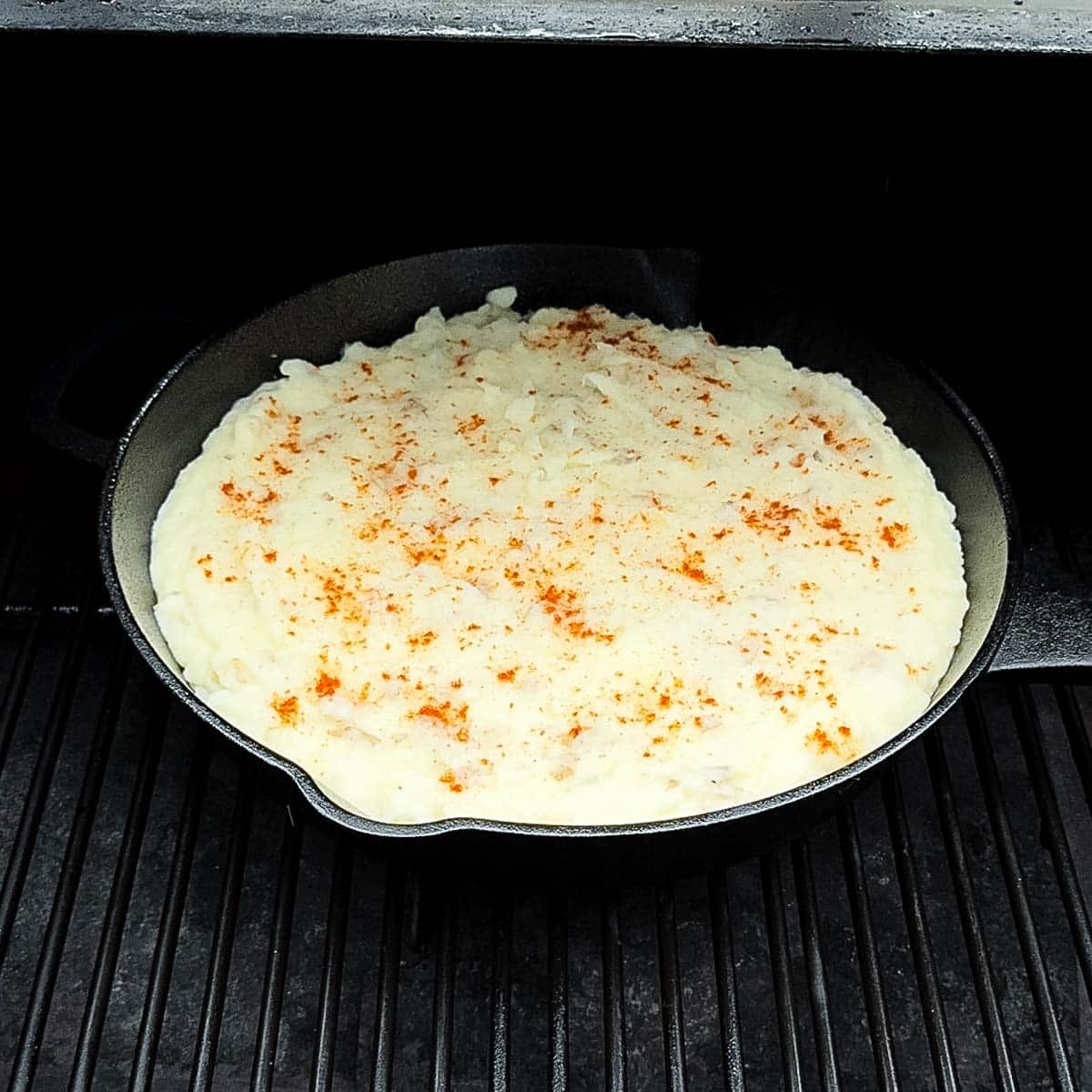 The skillet of mashed potatoes smoking on the grill grates of a pellet grill.
