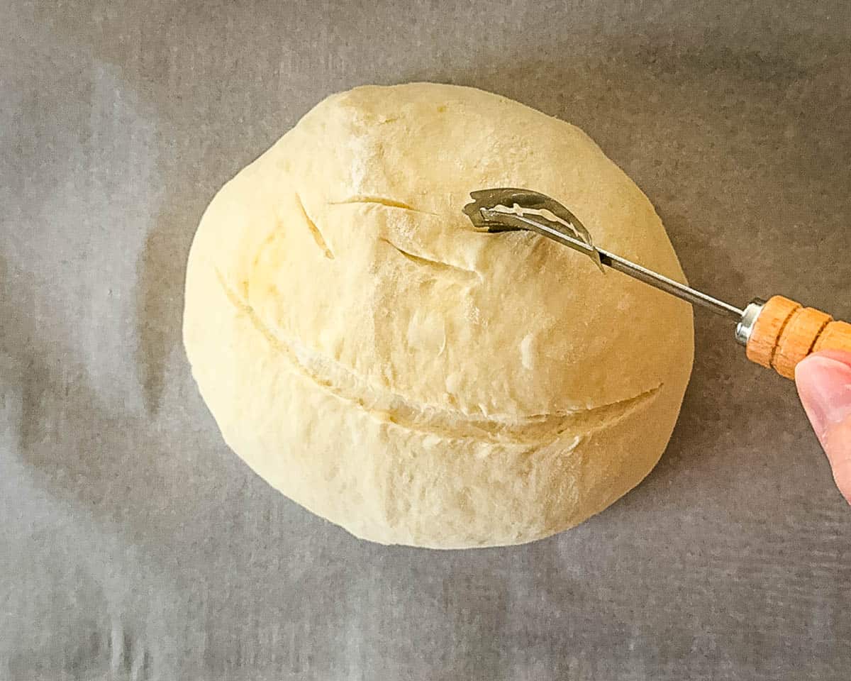 A bread lame scoring shallow slashes into the surface of the shaped sourdough discard dough.