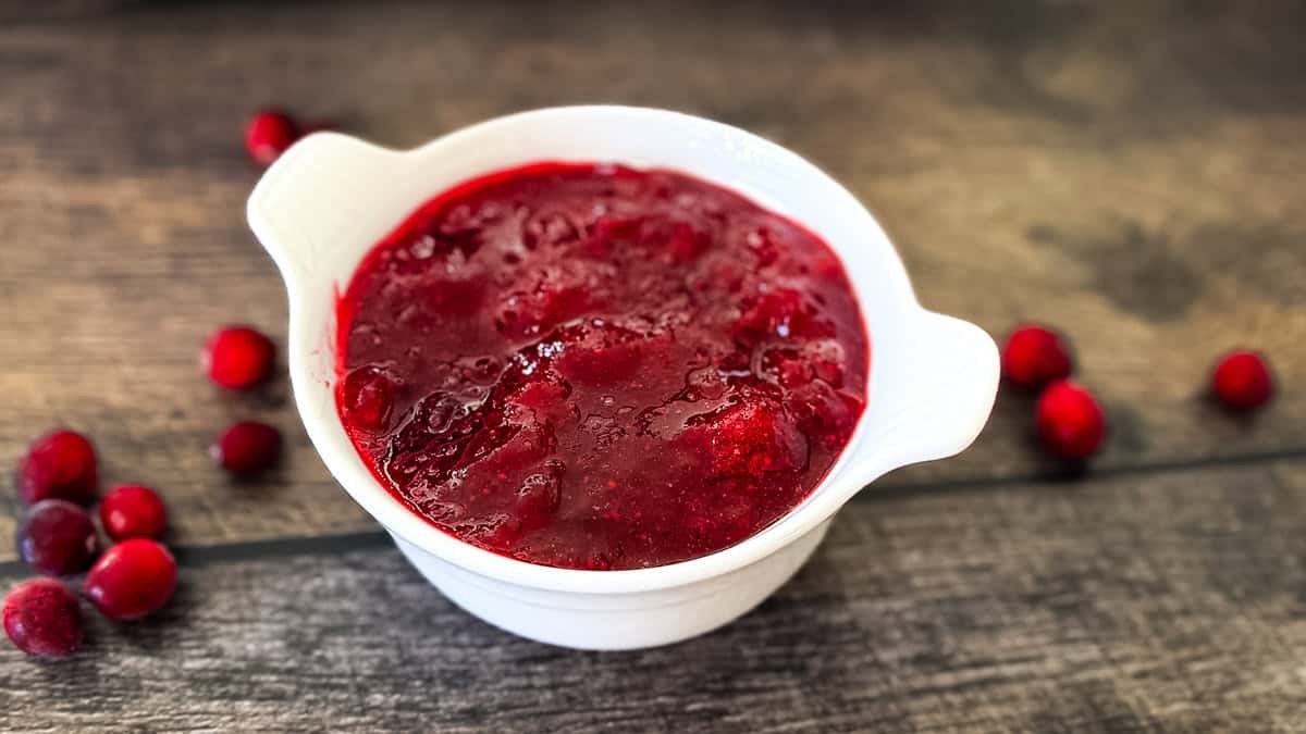 A white bowl filled with homemade cranberry sauce on a rustic wooden surface, surrounded by a few fresh cranberries.
