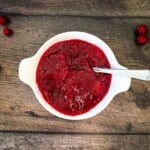 A white bowl filled with homemade cranberry sauce on a wooden table, surrounded by fresh cranberries.