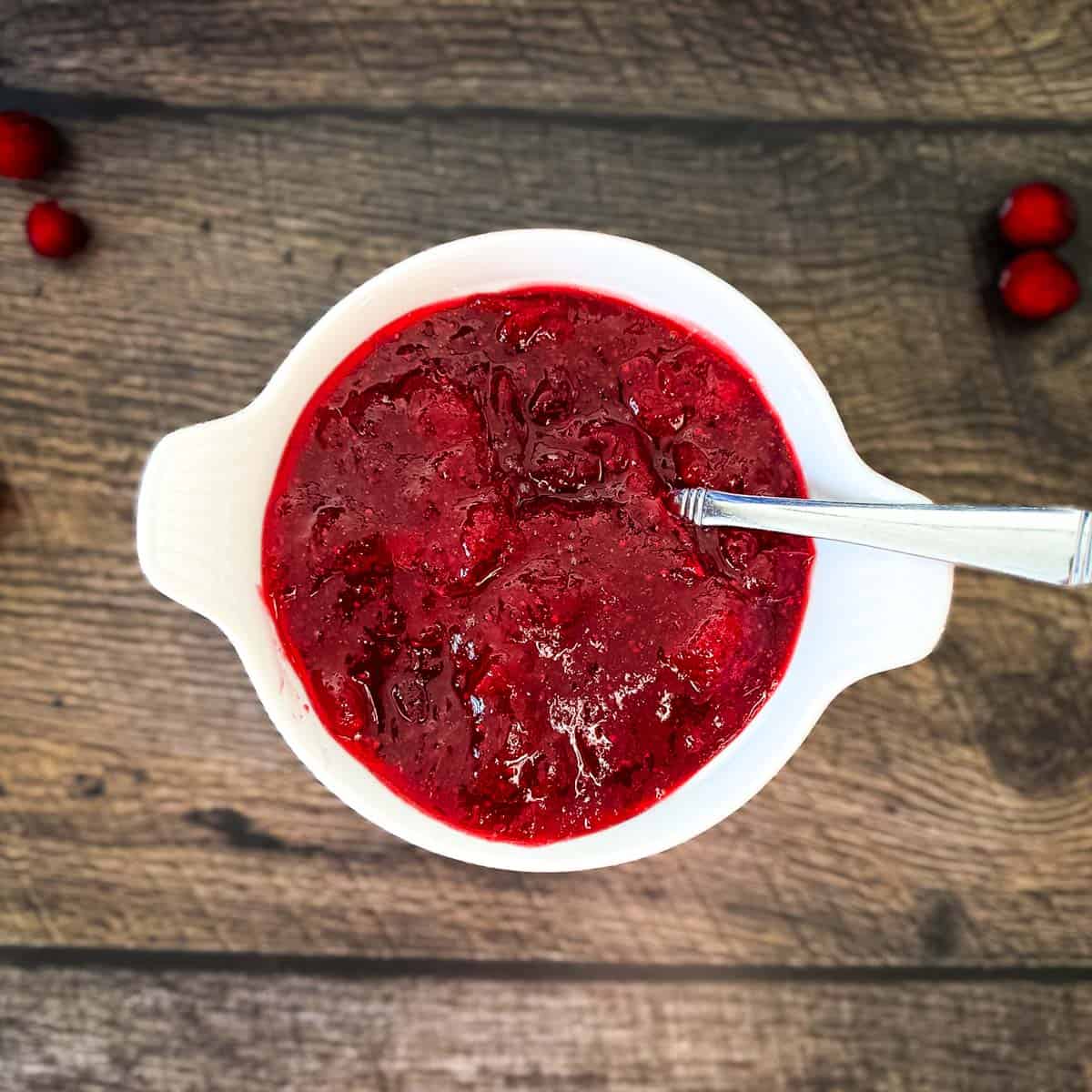 A white bowl filled with homemade cranberry sauce on a wooden table, surrounded by fresh cranberries.