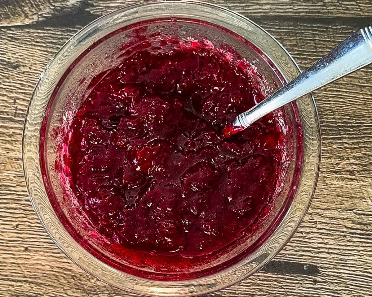 A glass bowl filled with thick homemade cranberry sauce, stirred with a silver spoon on a wooden table.