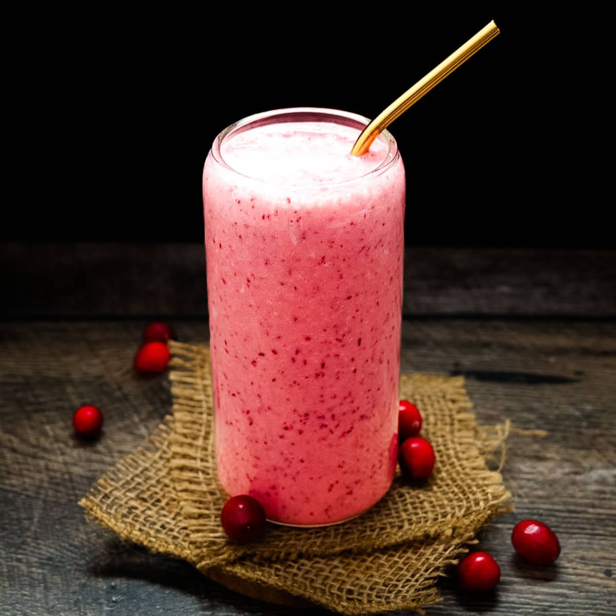 A tall glass of creamy pink cranberry smoothie with a gold straw, sitting on a rustic wood surface with fresh cranberries scattered around.