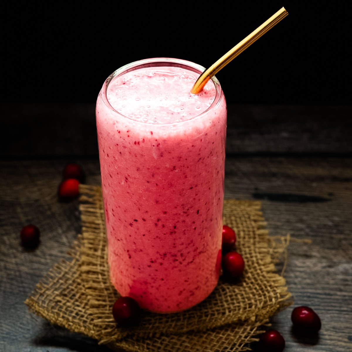 Cold and creamy cranberry smoothie in a clear glass with a gold straw, photographed on a dark wood table with cranberries around the base.