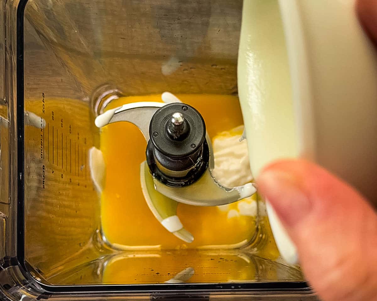 A hand pouring Greek yogurt into a blender filled with orange juice, showing the first step of building the cranberry smoothie base.