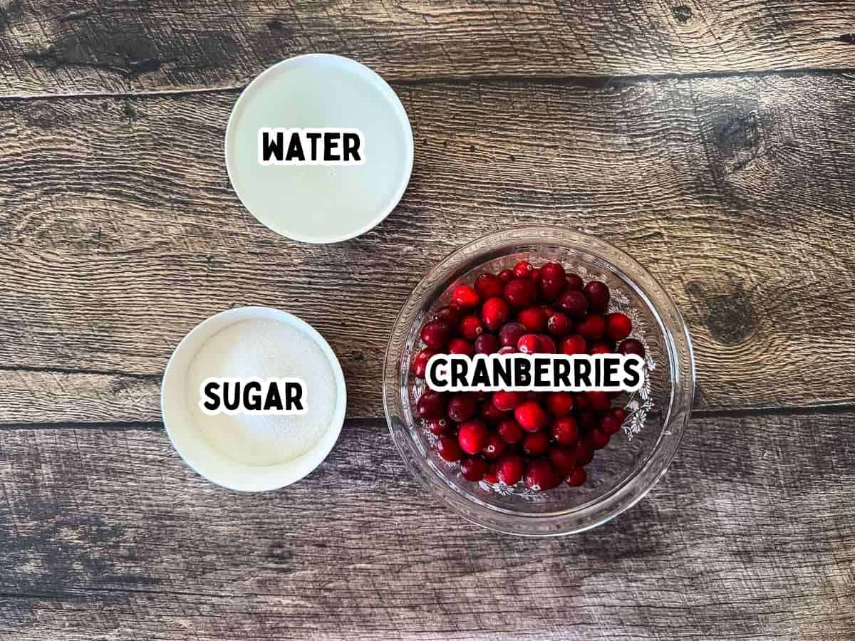 Three bowls on a wooden table showing the ingredients for homemade cranberry sauce — fresh cranberries, sugar, and water labeled with text.