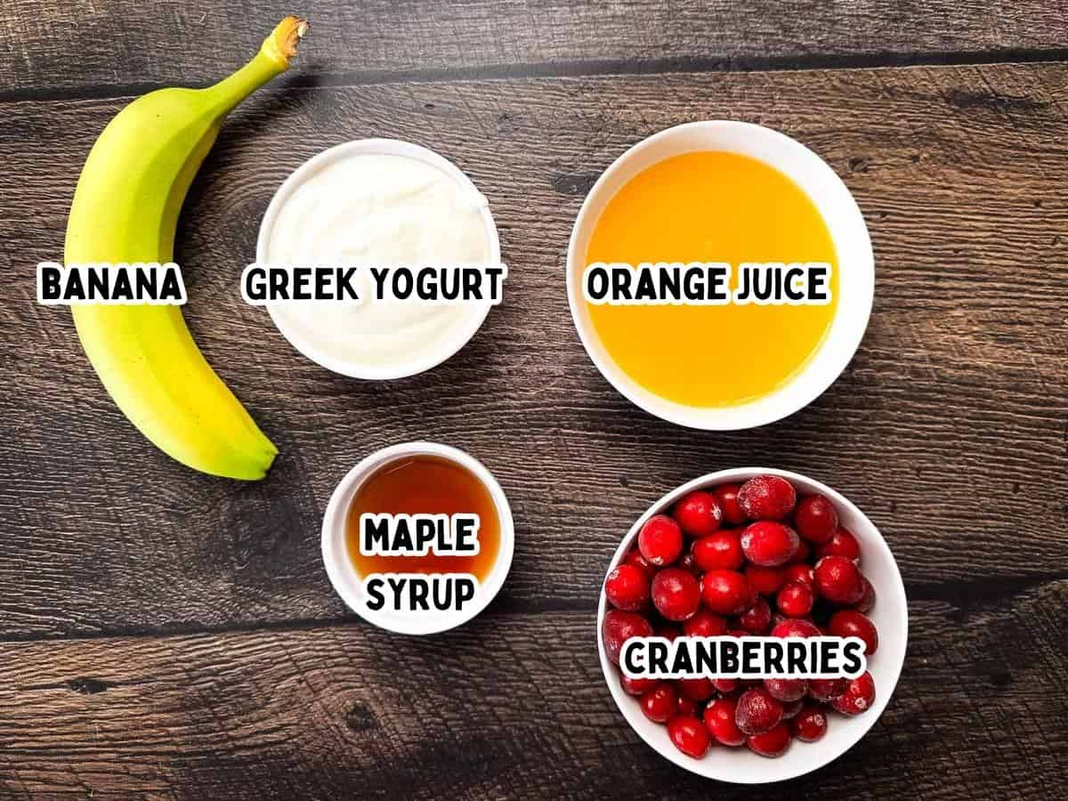 Five simple ingredients laid out on a wooden surface: a banana, a bowl of Greek yogurt, a bowl of orange juice, a small dish of maple syrup, and a bowl of fresh cranberries.
