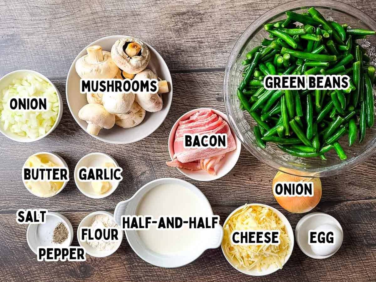 Labeled ingredients for smoked green bean casserole laid out on a wooden surface, including green beans, mushrooms, onions, bacon, butter, garlic, flour, half-and-half, cheese, egg, salt, and pepper.
