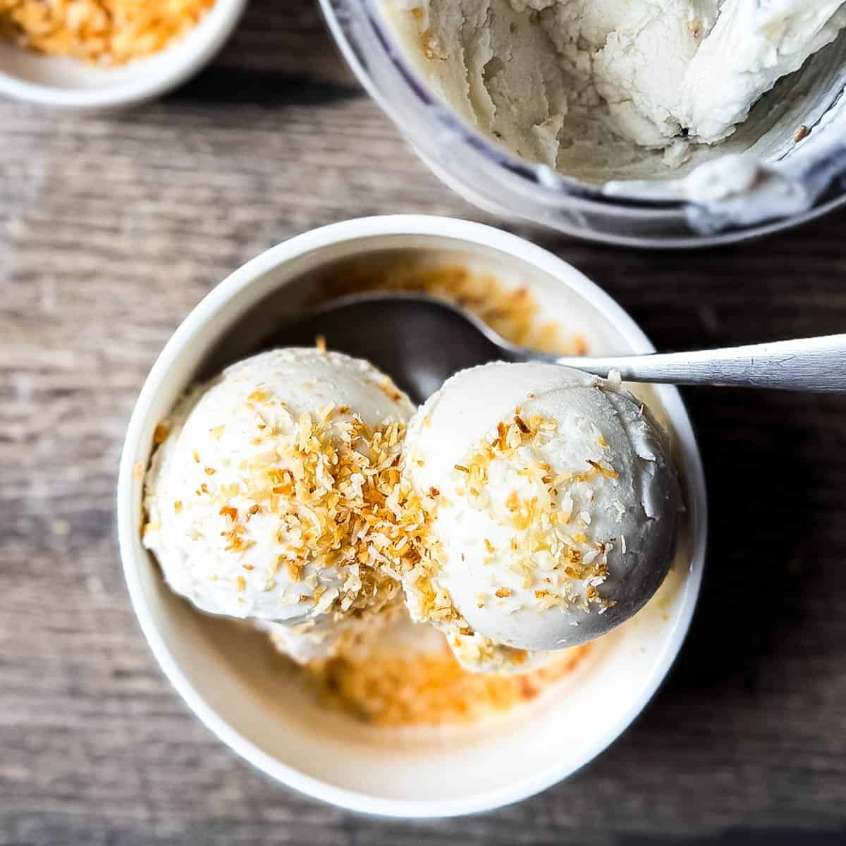 Overhead view of two scoops of Ninja Creami coconut ice cream sprinkled with golden toasted coconut flakes in a white bowl on a wooden table.