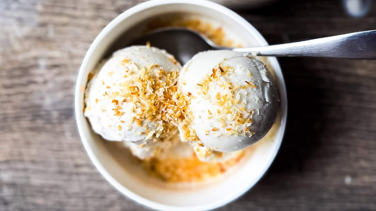Overhead view of two scoops of Ninja Creami coconut ice cream sprinkled with golden toasted coconut flakes in a white bowl on a wooden table.