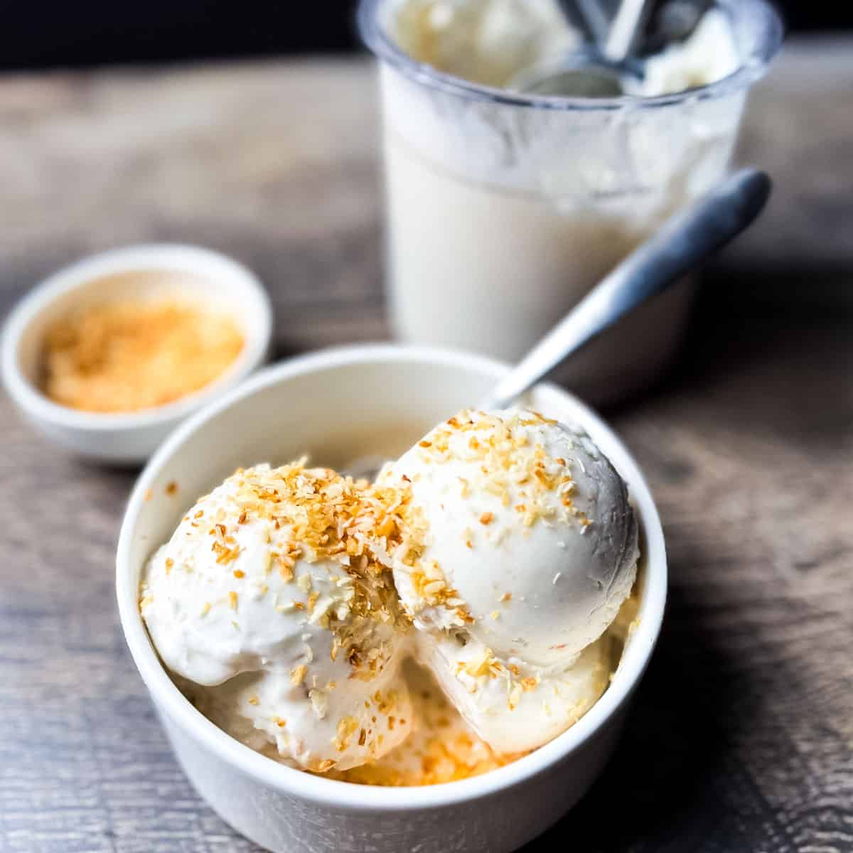 Bowl of Ninja Creami coconut ice cream on a rustic wooden surface with the Creami pint and toasted coconut in the background.