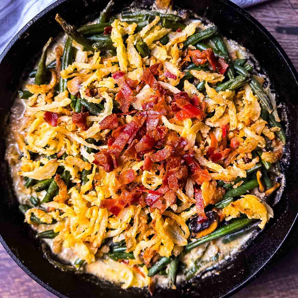 Smoked green bean casserole in a cast iron skillet topped with crispy onions and smoky bacon pieces.