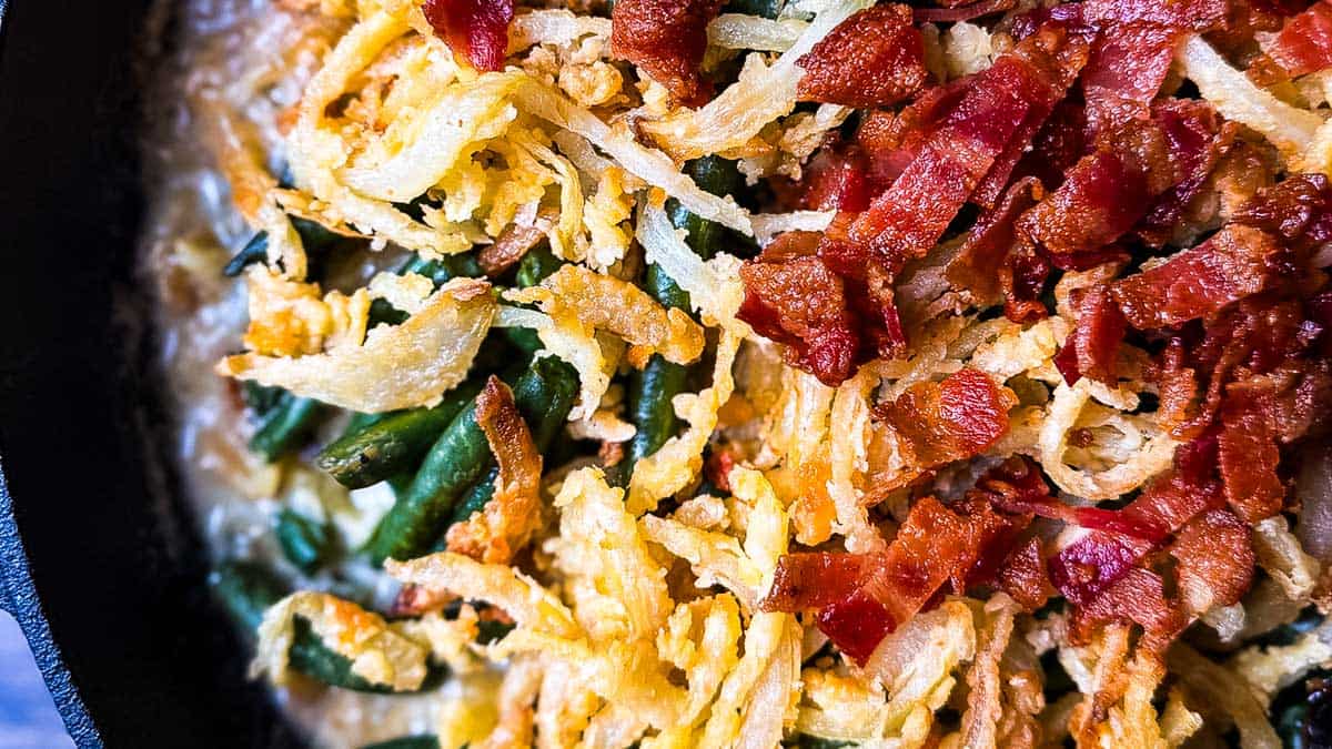 Smoked green bean casserole topped with golden onions and chopped bacon in a cast iron skillet.