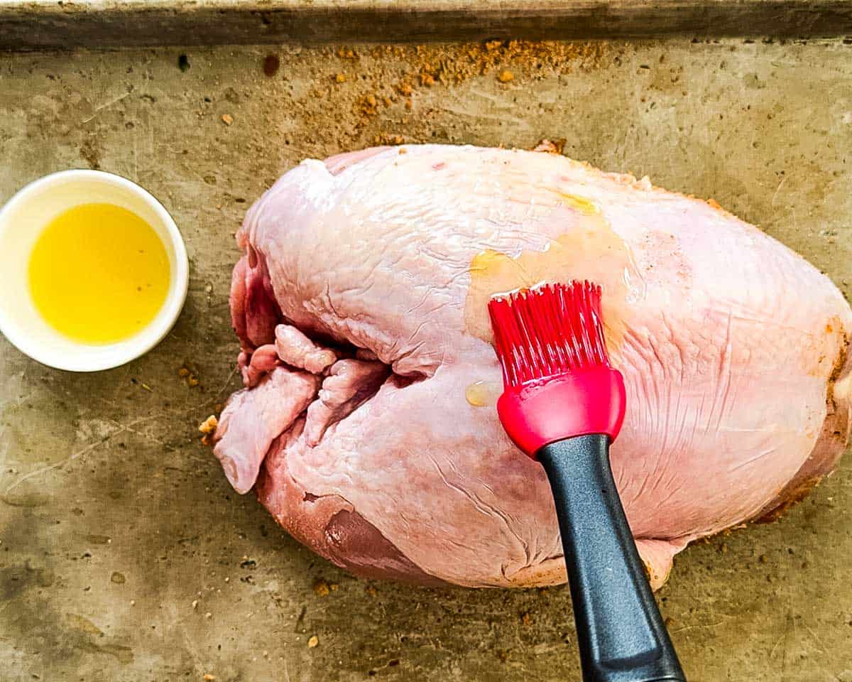 Uncooked turkey breast being brushed with olive oil using a red silicone brush, with a small bowl of oil beside it on a baking sheet.
