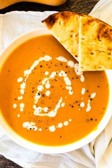 Bowl of roasted butternut squash soup with a swirl of cream, cracked black pepper, and warm flatbread on the side.