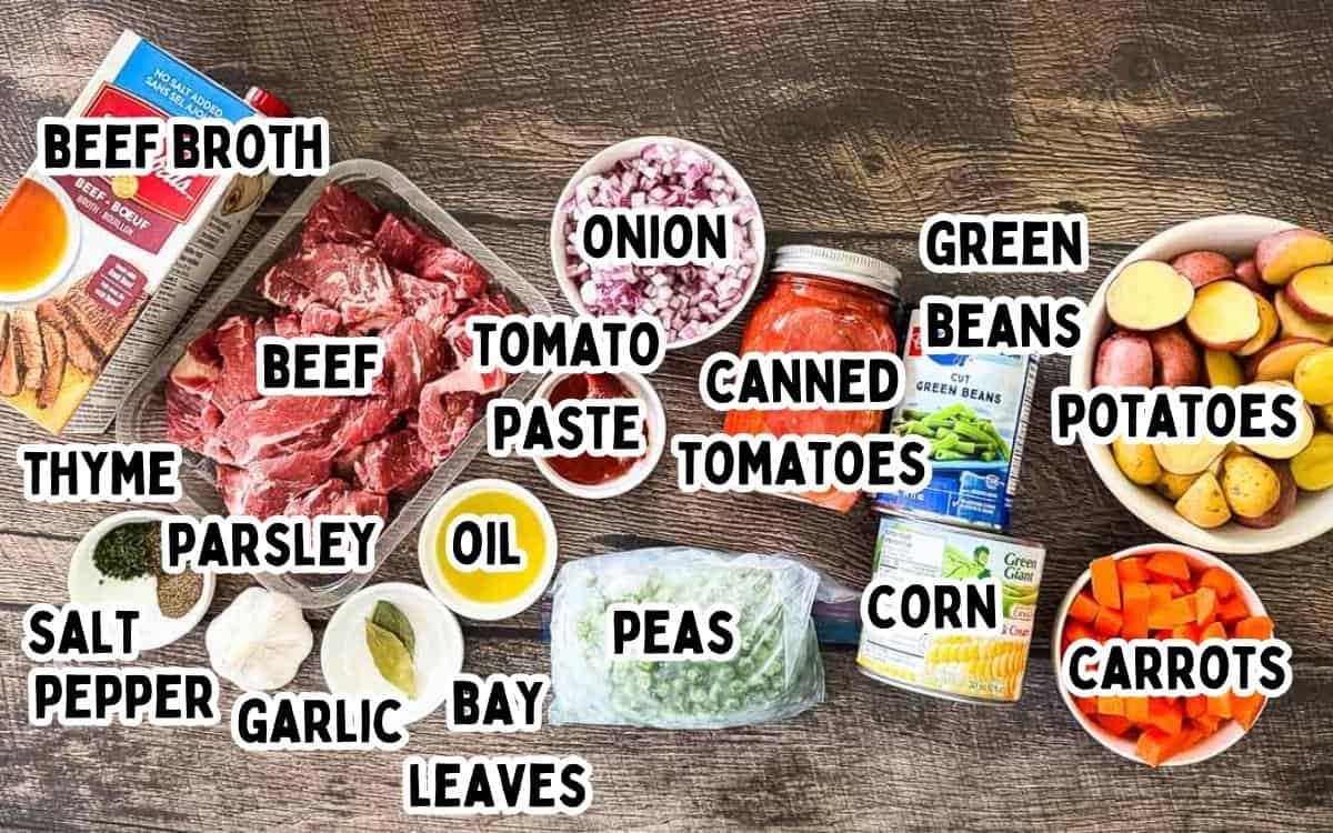 Ingredients for old-fashioned beef vegetable soup including beef chuck, potatoes, carrots, green beans, peas, corn, tomatoes, onion, garlic, and seasonings laid out on a wooden surface.