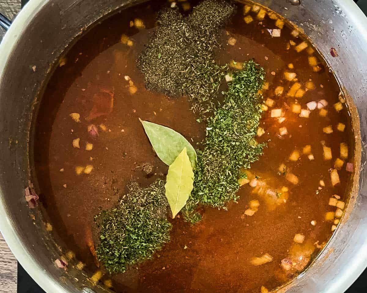 Beef broth, diced tomatoes, herbs, and bay leaves added to the pot for simmering beef vegetable soup.